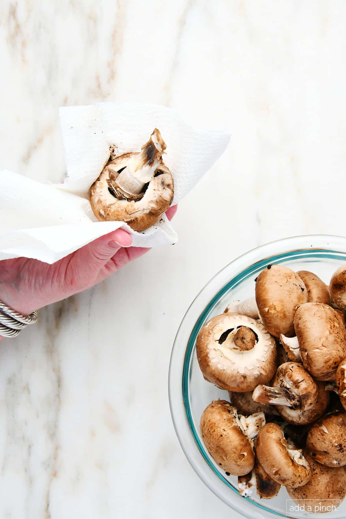 Fresh whole mushrooms in a glass bowl with one being cleaned with a damp paper towel.