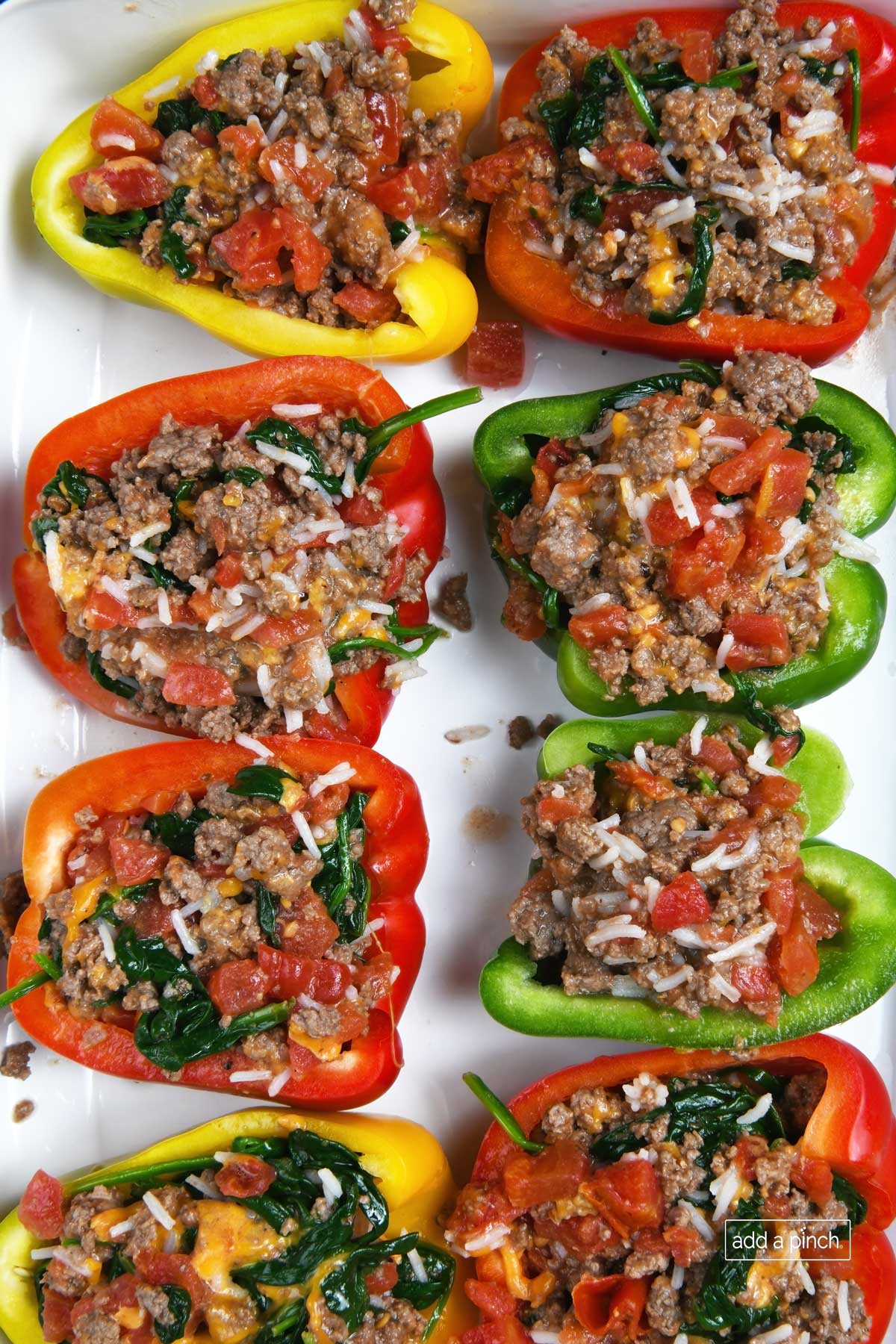 Colorful bell pepper halves are stuffed with meat, rice, spinach, tomato and cheese, ready to bake in a white baking dish.