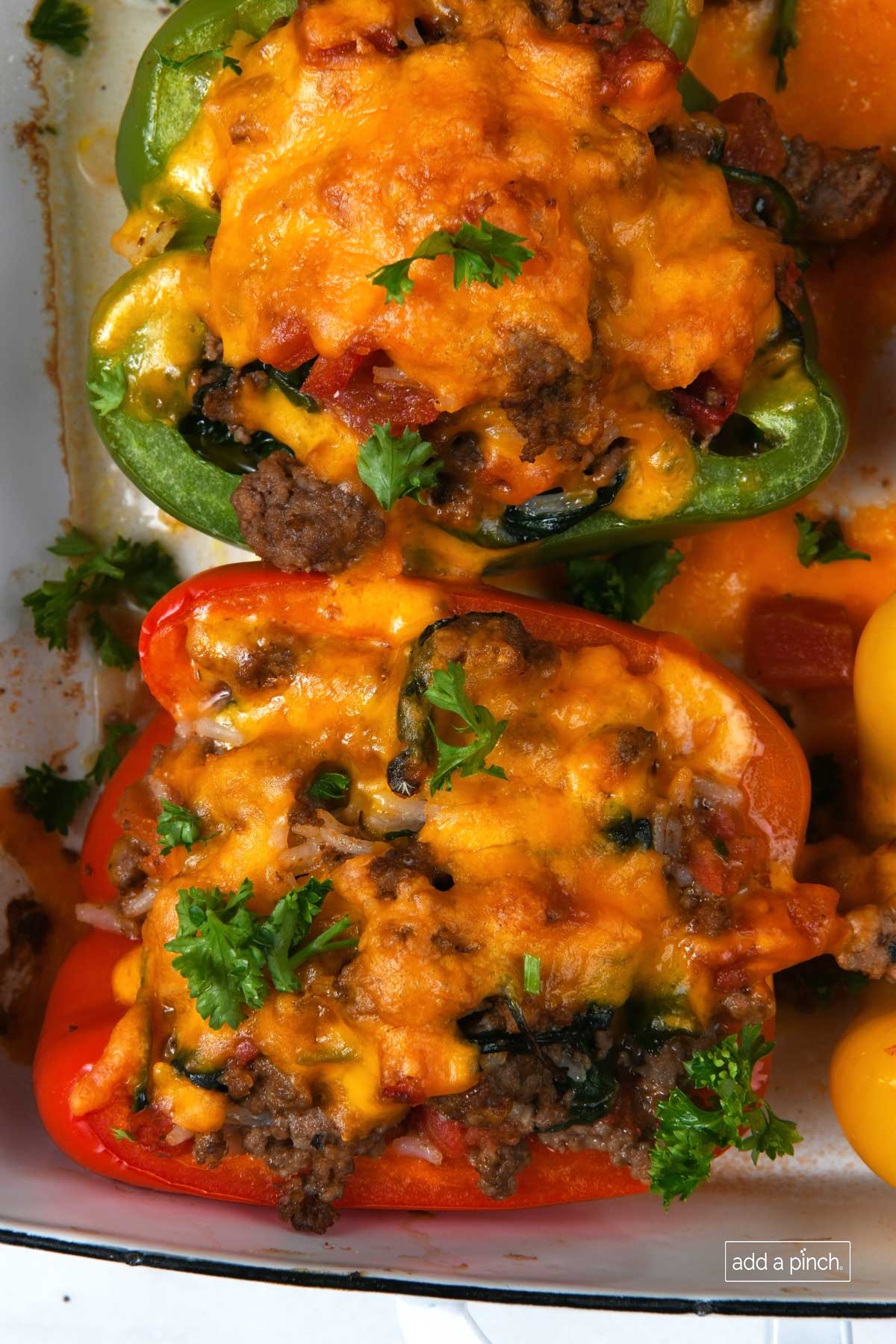 Photo of red, green and yellow peppers halved and filled with a beef and rice mixture and topped with cheddar cheese in a white baking dish.