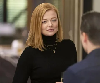 A woman with red hair in a black turtleneck, wearing a necklace, holds a glass while talking to a man in a suit.