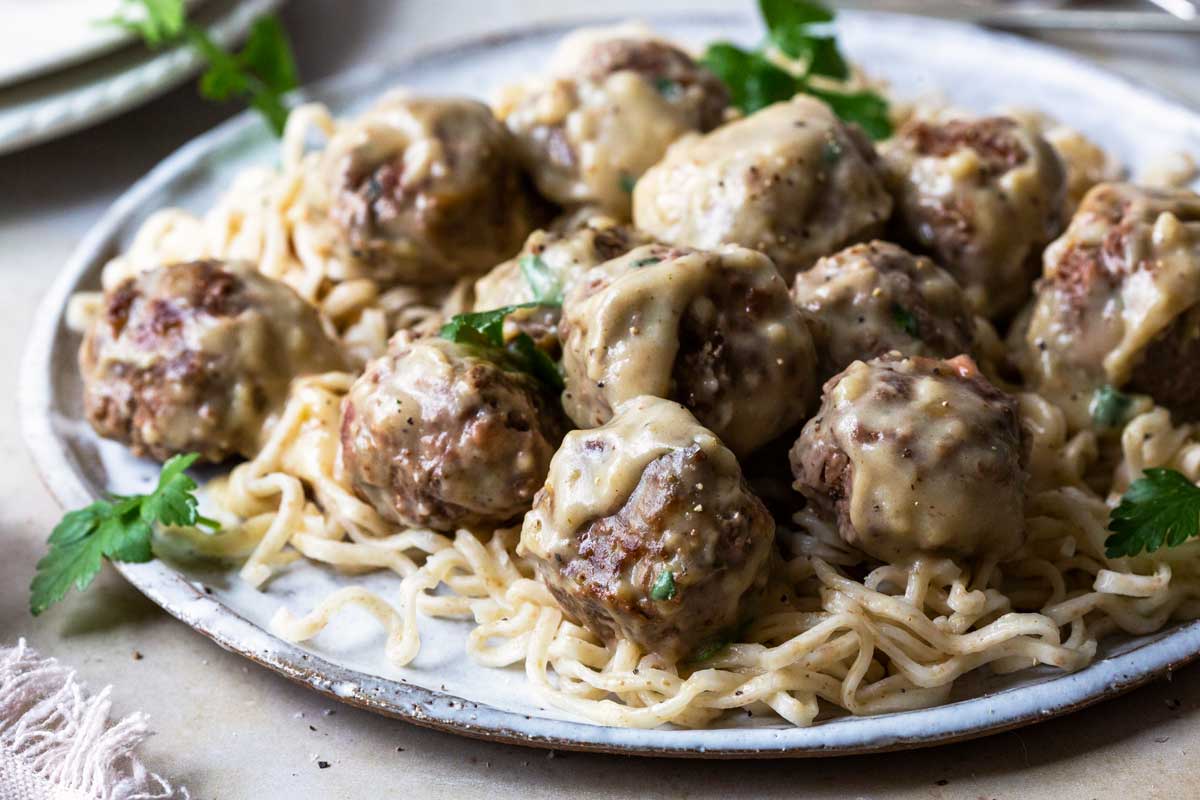 Photograph of Swedish Meatballs covered in sauce atop noodles on a white plate garnished with parsley.