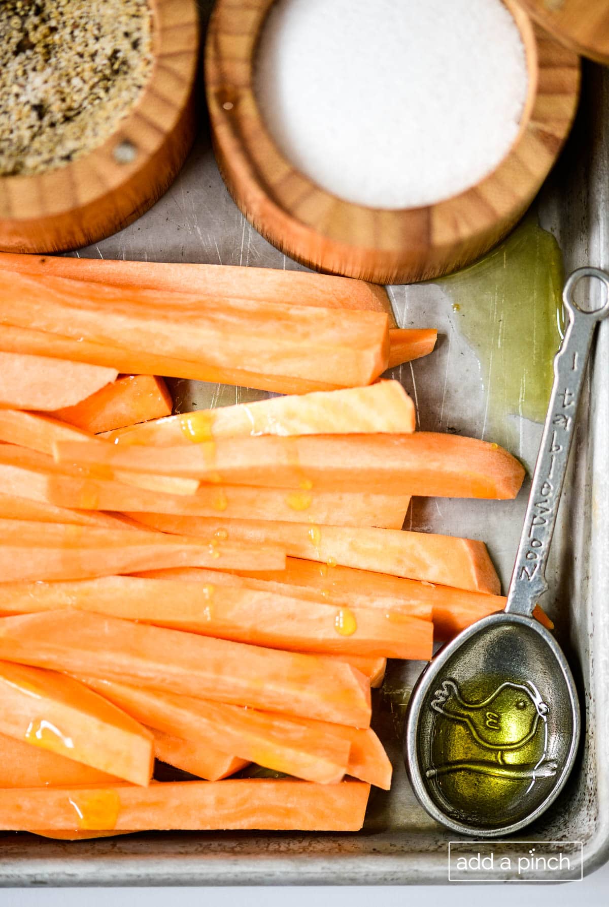 Photograph of sweet potato fries, olive oil and salt on a baking sheet with a measuring spoon. 