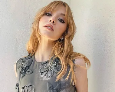 A woman with long blonde hair wearing a floral-patterned dress poses against a pale background.