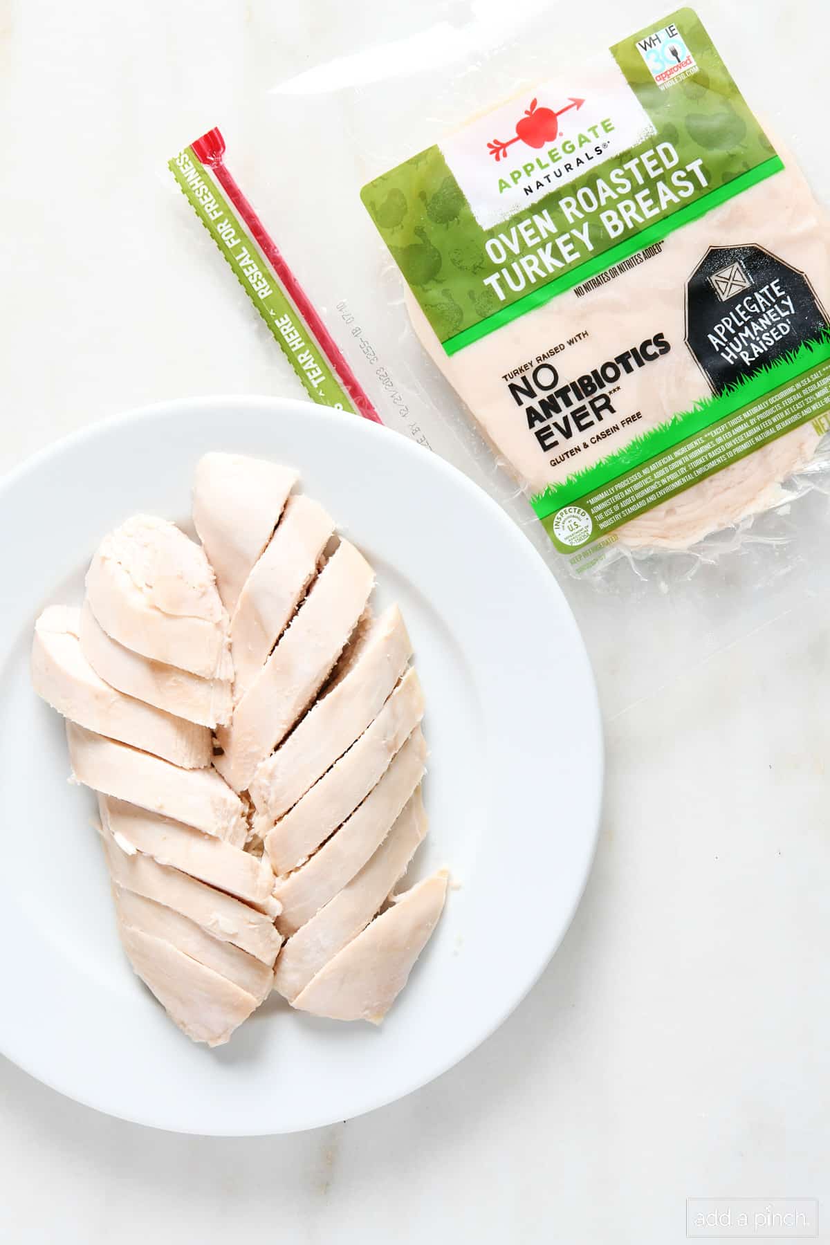 Sliced turkey breast on a white plate and package of sliced turkey breast on marble counter.