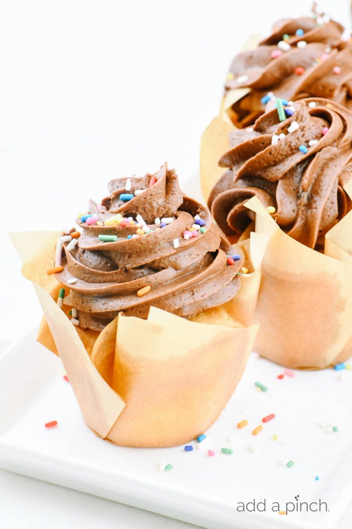 A white cake plate holds vanilla cupcakes frosted with chocolate buttercream and sprinkles, wrapped in parchment.