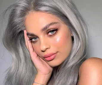 Woman with long silver-gray hair and elegant makeup, resting her face on her hand, gazing into the camera.